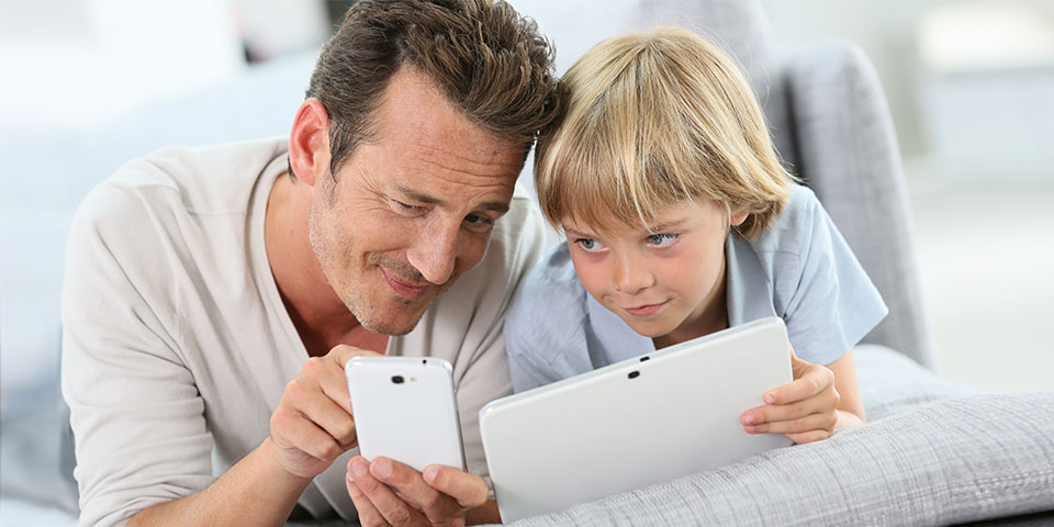 Vater und Sohn liegen auf Sofa und lesen am Handy und Tablet