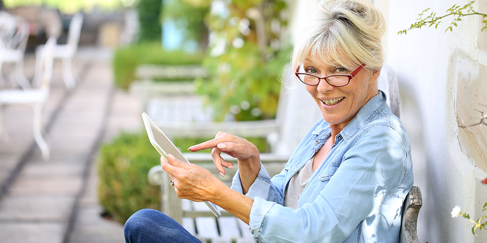 Ältere Frau mit Brille sitzt auf Terrasse und liest auf einem Tablet